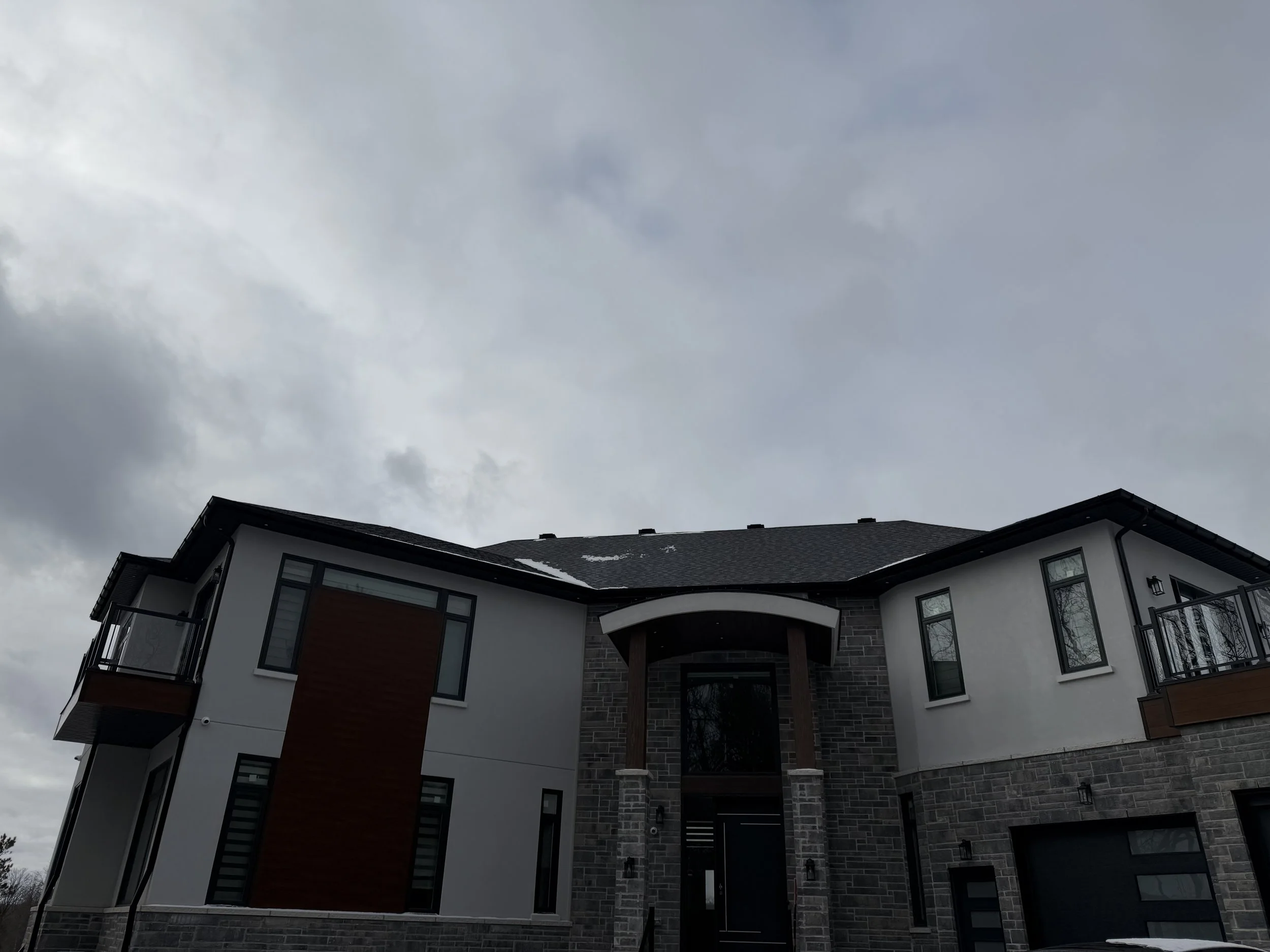 Modern multi-story house with dark roof, white walls, brown accents, and large windows, under cloudy sky.