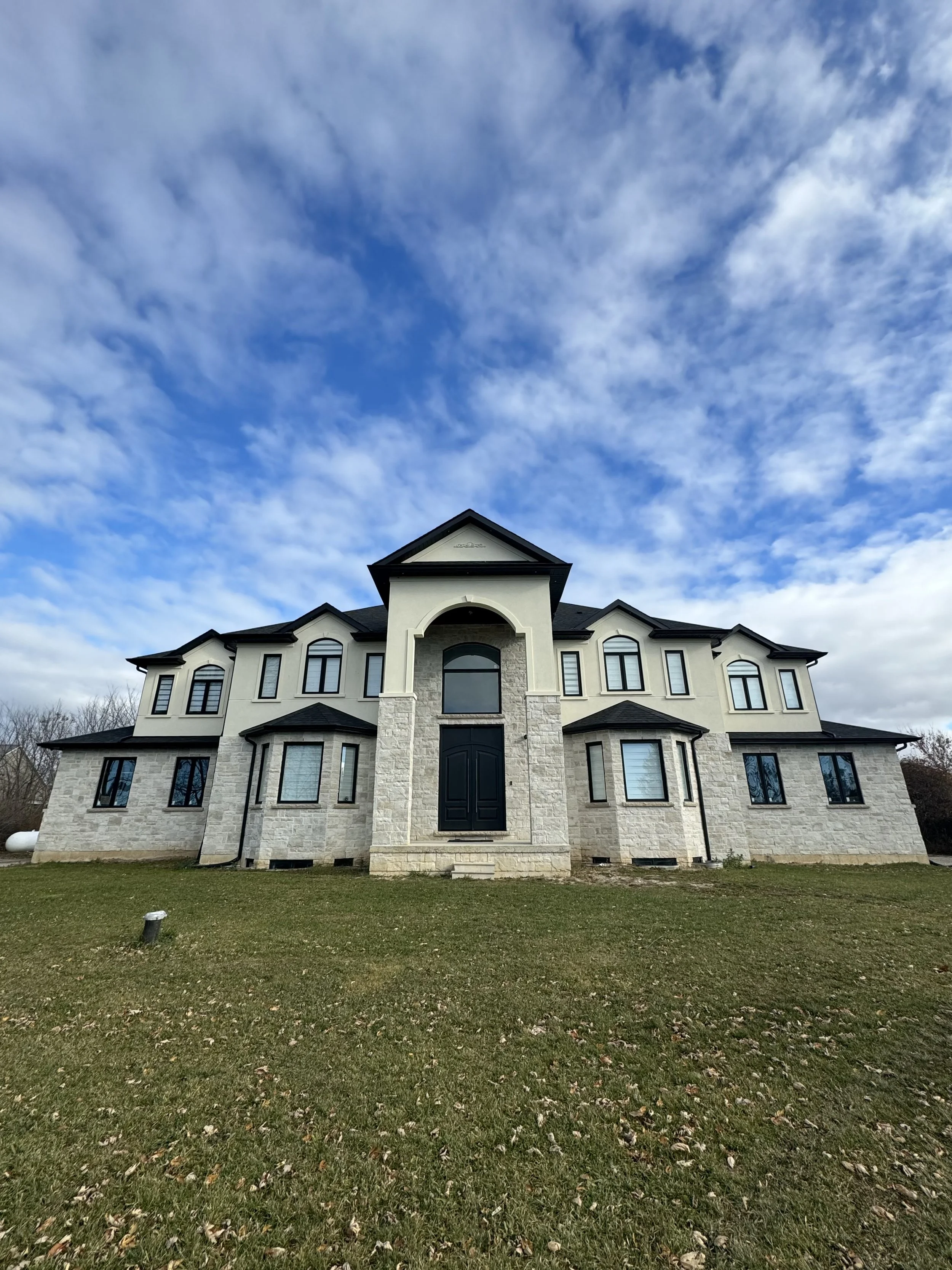 Large modern house with white and gray stone exterior, multiple windows, and a front entrance with a black door, set against a backdrop of a partly cloudy sky and a grassy lawn.