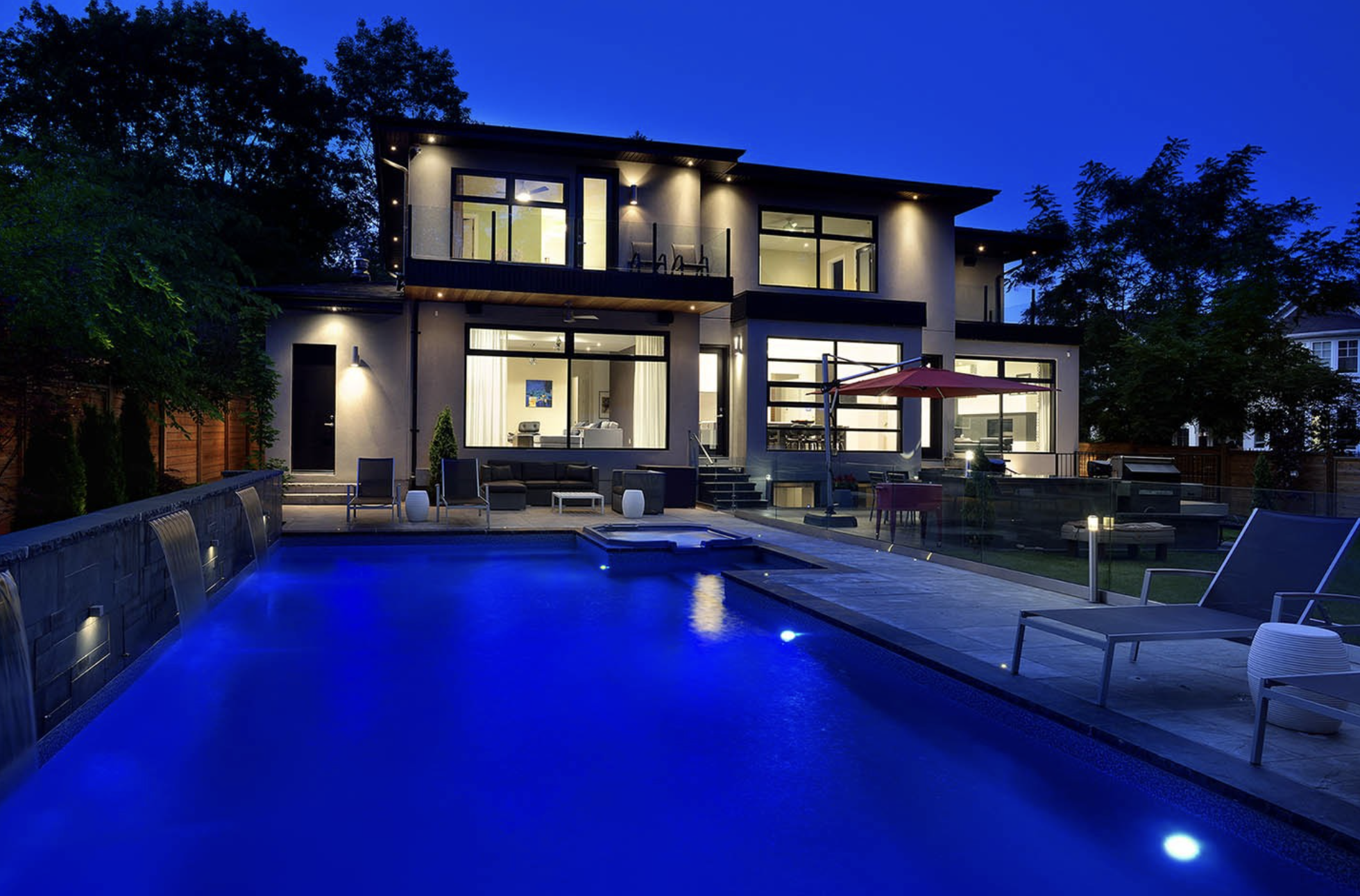 Modern house with pool at night, illuminated outdoor lighting, outdoor furniture, and a fence surrounding the yard.