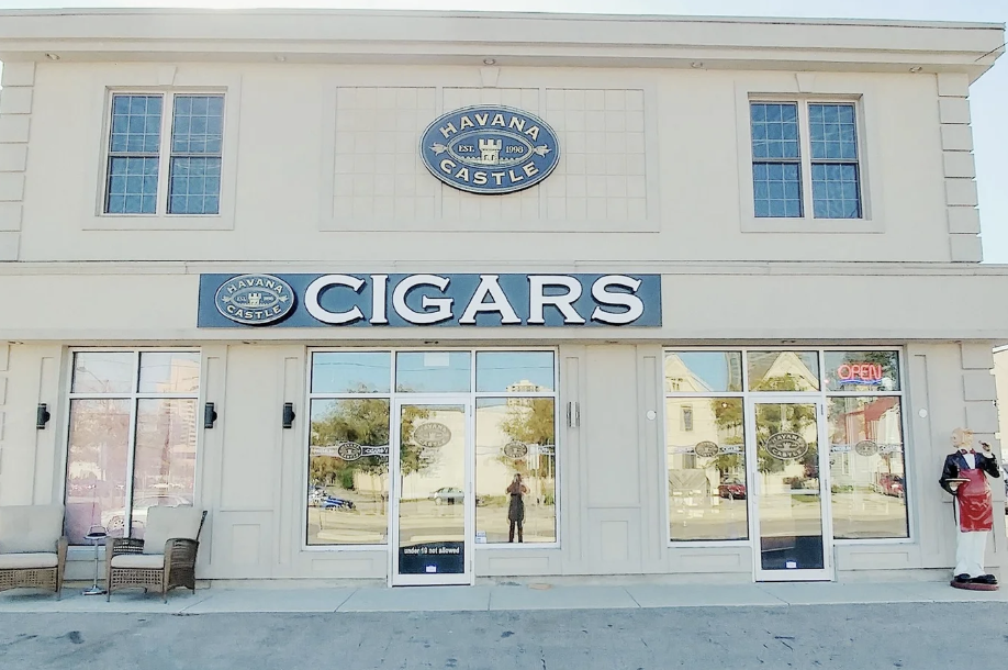 Front of a cigar shop called Havana Castle with large windows, a sign that says 'Cigars,' and a statue of a man in a red apron holding a cigar outside.