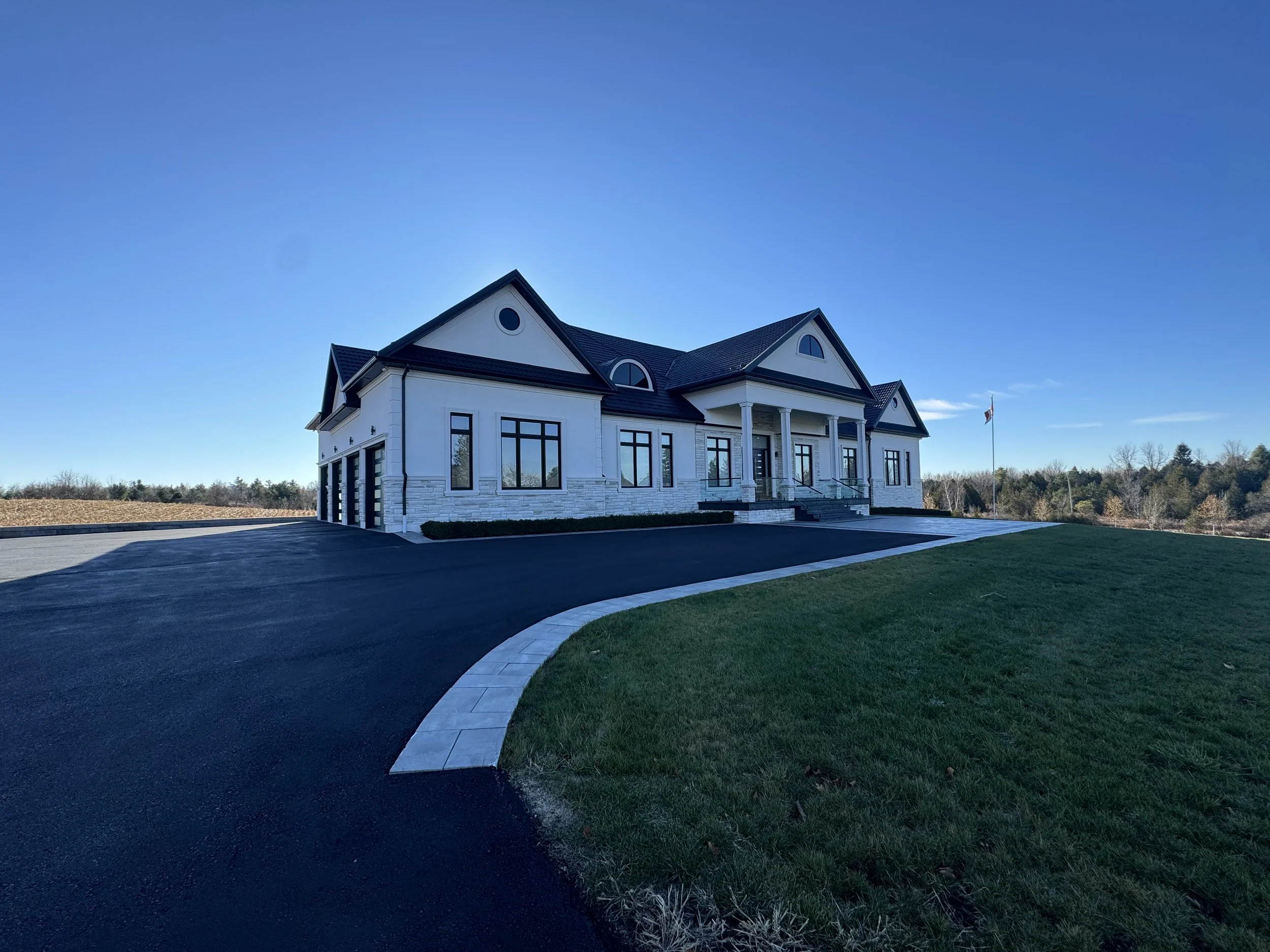 Large white house with black roof and multiple windows, surrounded by green lawn and asphalt driveway, under a clear blue sky.