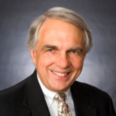 Portrait of a middle-aged man with gray hair, wearing a black suit and patterned tie, smiling against a gray background.