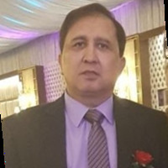 A man in a dark suit with a purple shirt and striped tie, standing indoors with purple lighting and curtains in the background.