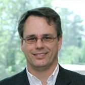 A man with dark hair, glasses, and a suit smiling at the camera, with a background of trees and windows.