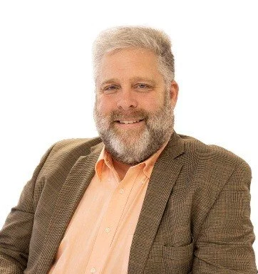 A smiling middle-aged man with gray hair and beard, wearing a brown blazer and peach shirt, sitting against a white background.