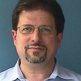 A man with glasses, dark hair, and a goatee, wearing a light blue shirt, standing against a blue background.
