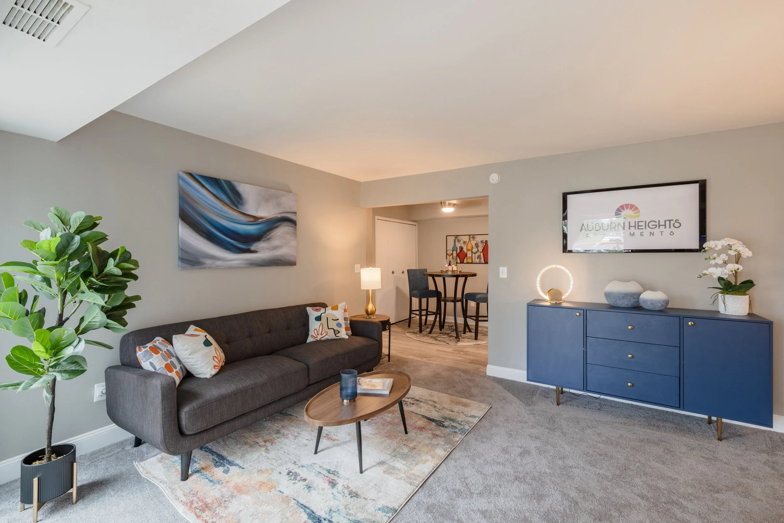 Living room with a gray sofa, colorful pillows, a patterned rug, a wooden coffee table, abstract wall art, a potted plant, a blue sideboard with decorative objects, a wall-mounted TV, and a small dining area in the background with high tables and chairs.