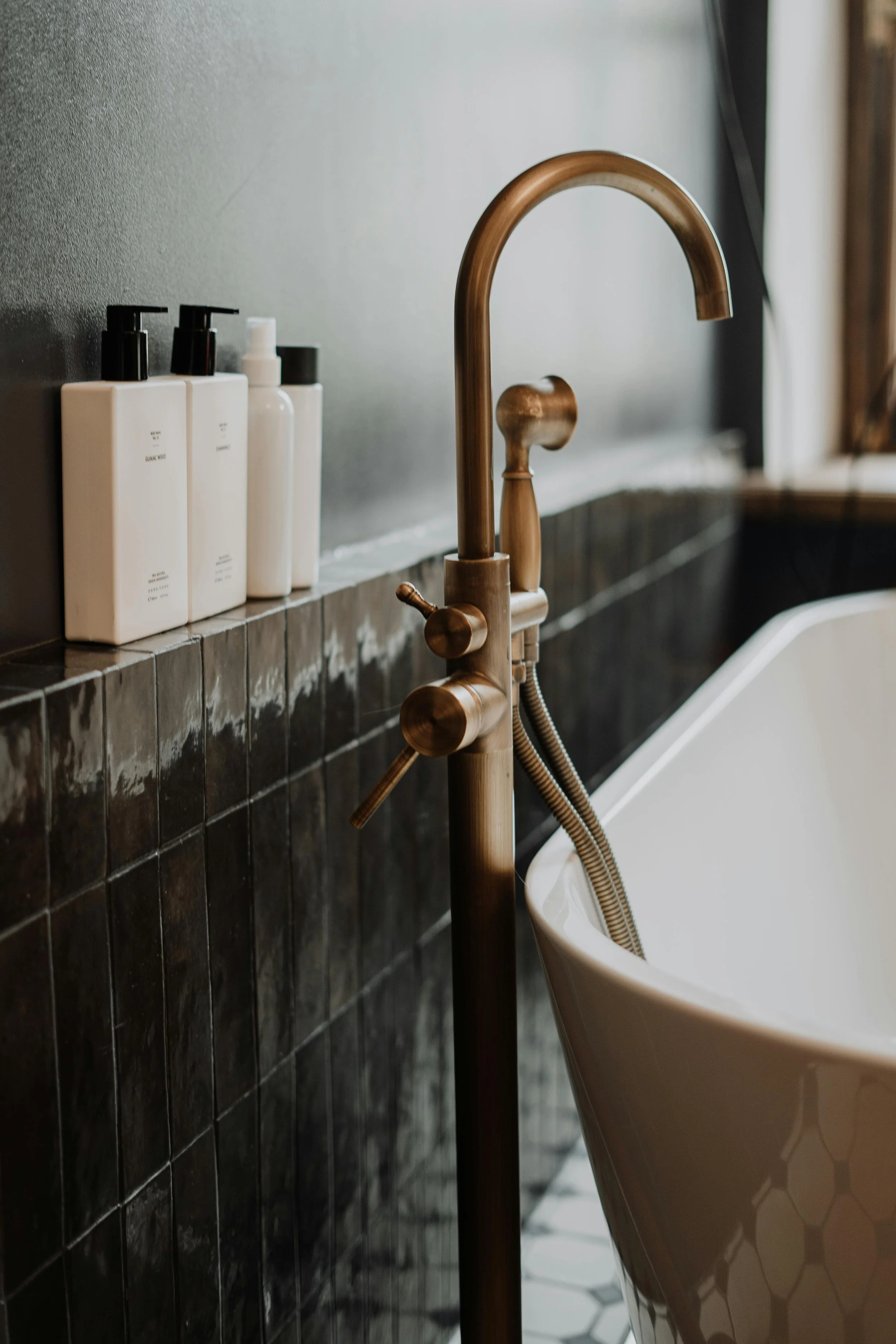 Close-up of a modern bathtub faucet with a handheld shower and a dark tiled wall in a bathroom.