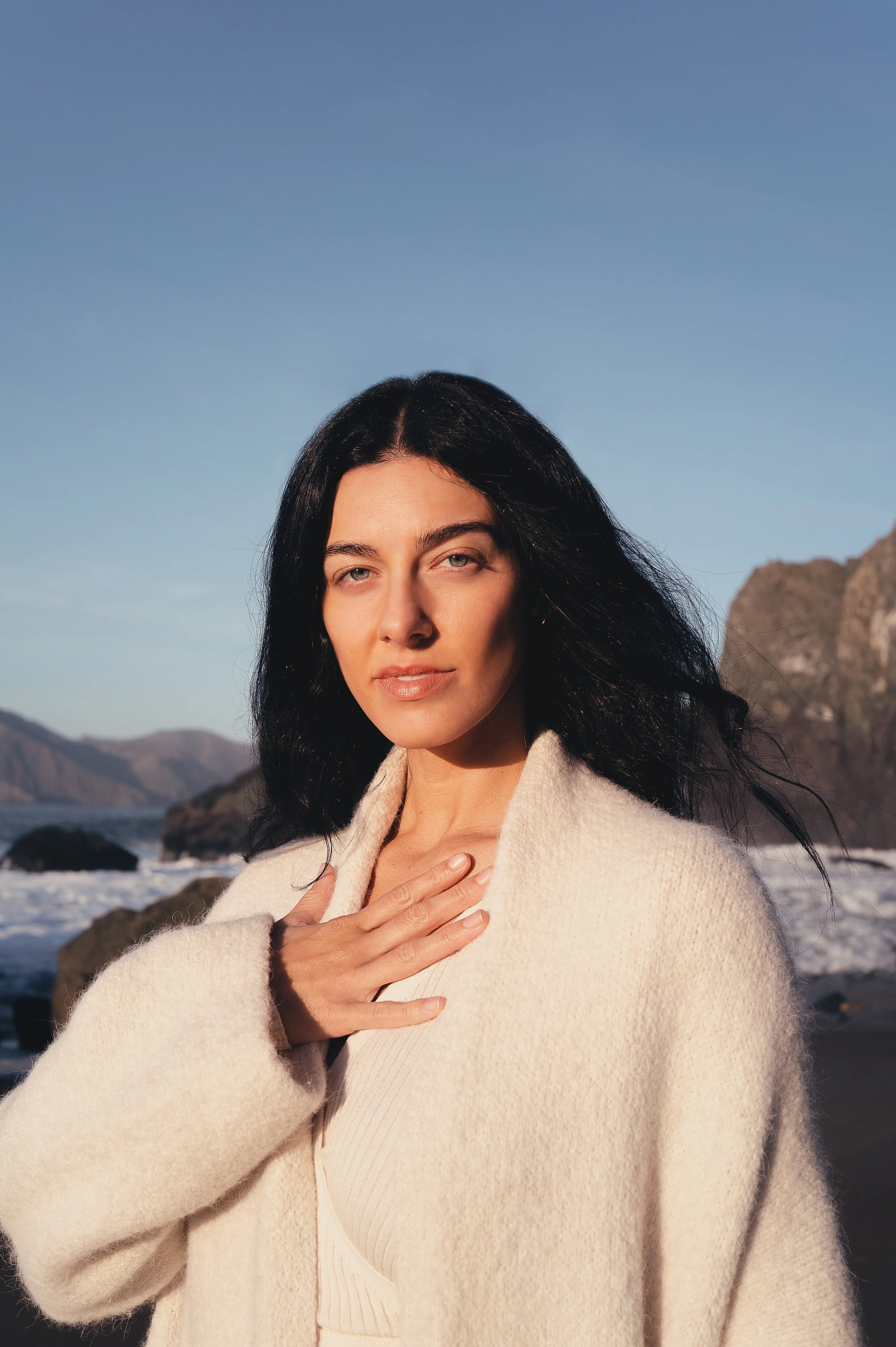A woman with long dark hair standing on a beach with rocks and mountains in the background during sunset.