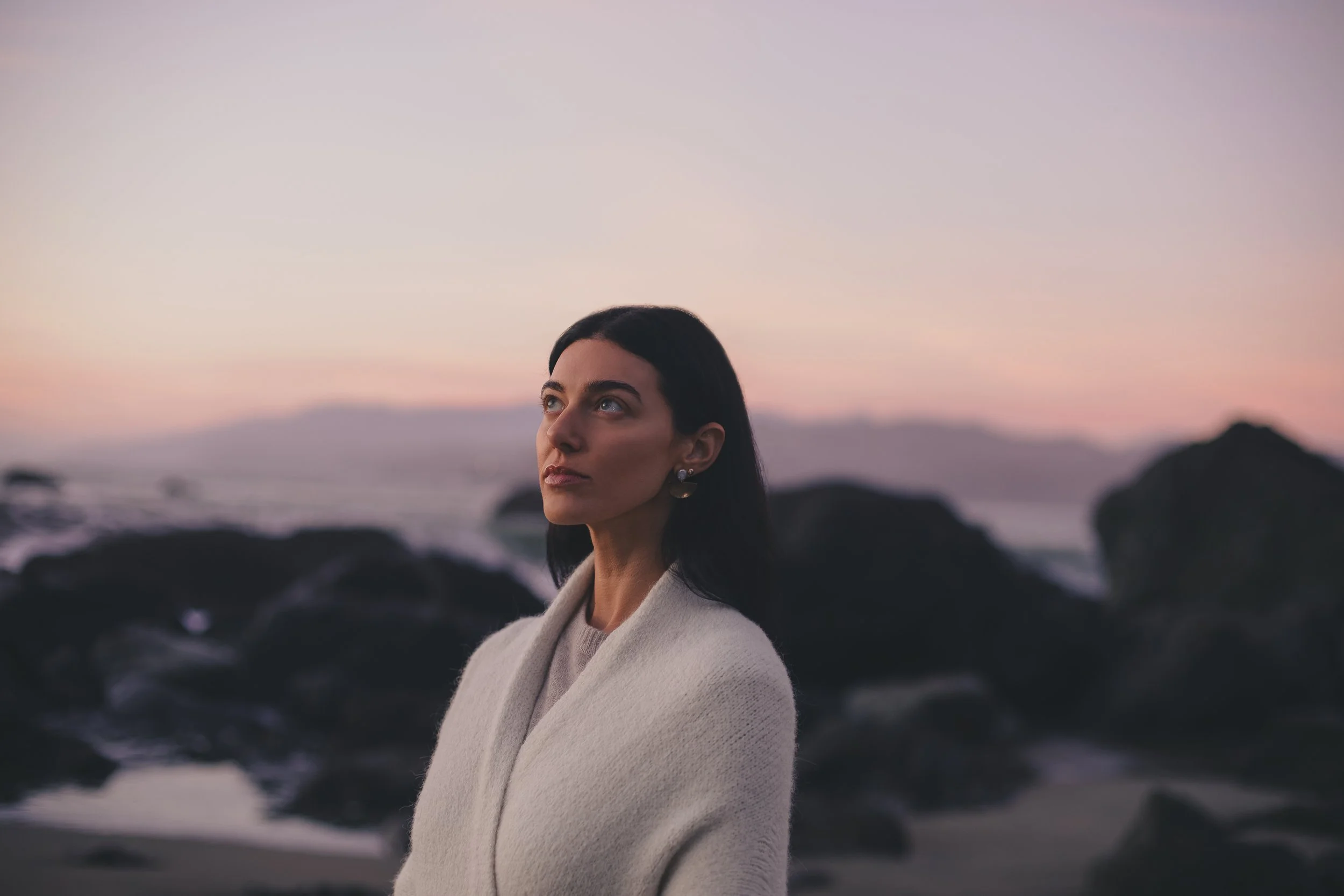 A woman with dark hair and earrings stands on a rocky beach at sunset, looking thoughtfully into the distance.