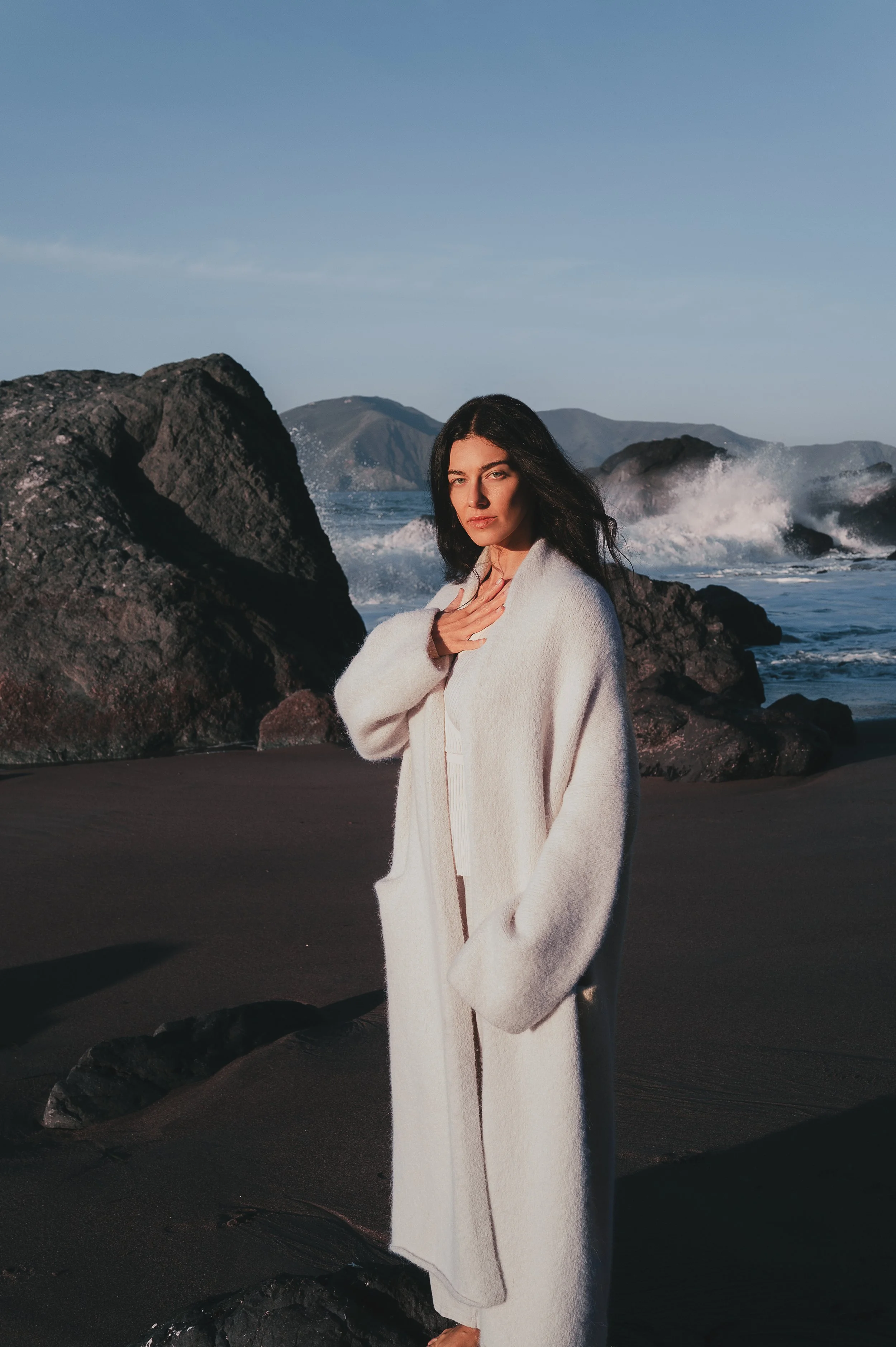 Woman standing on dark sandy beach near rocks, ocean waves crashing behind her, wearing a long white coat, with mountains in the background.