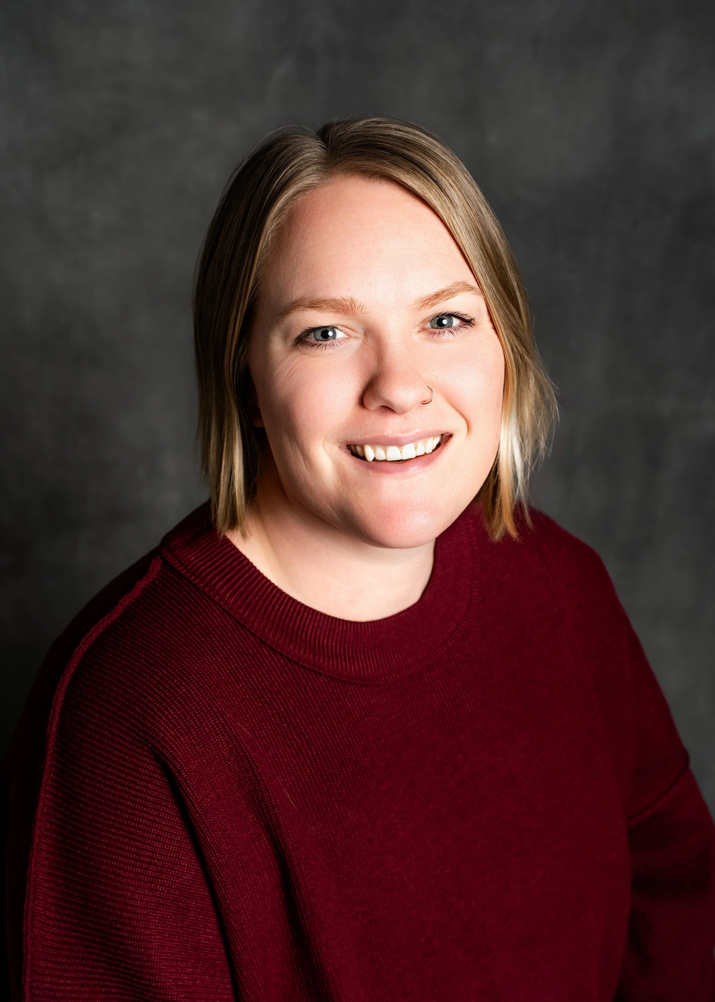 Portrait of teacher Ms.Kelly smiling. Woman with long, blonde hair, wearing a pink textured top, against a dark gray background.