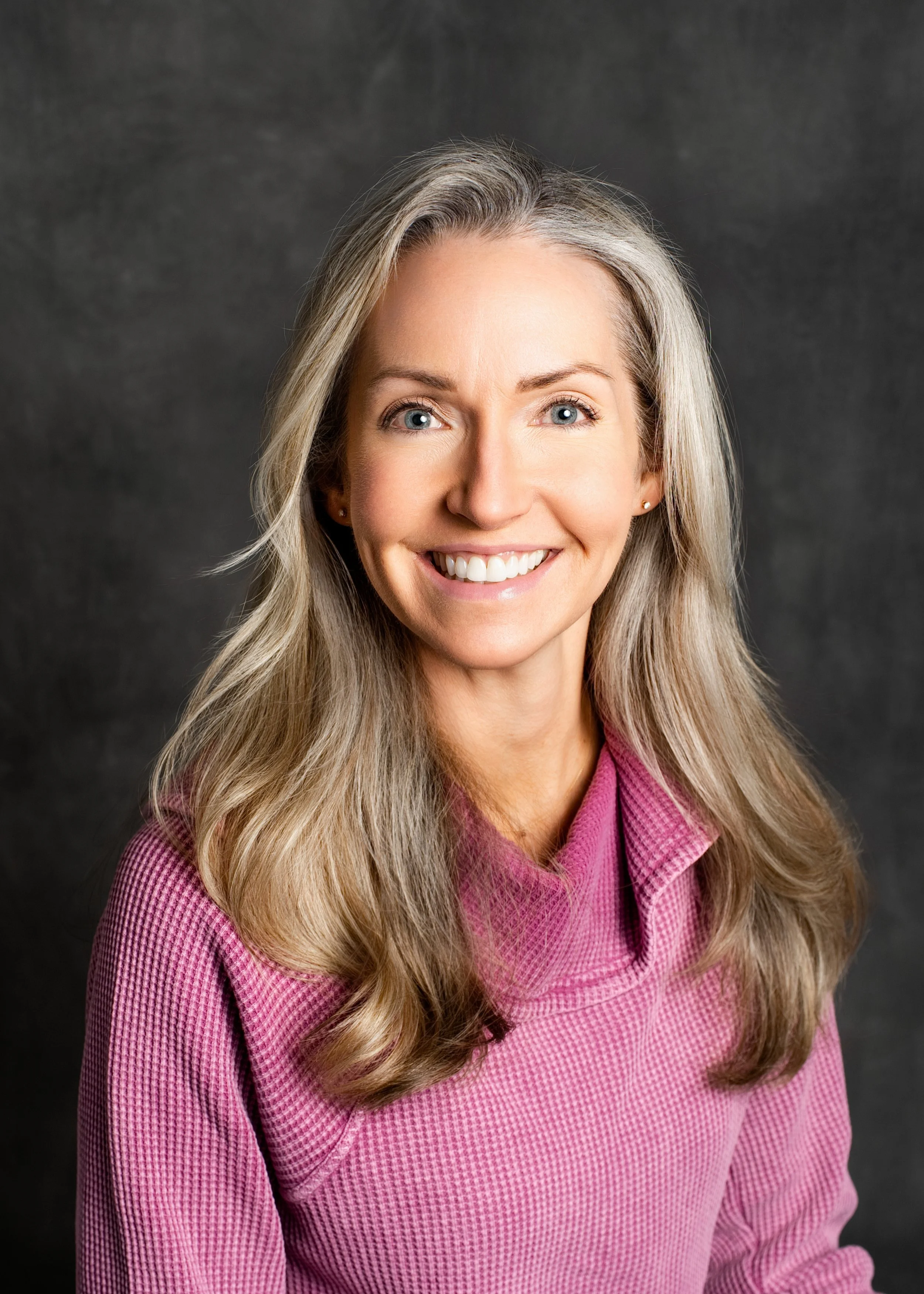 Portrait of teacher Ms.Jennifer smiling. Woman with long, blonde hair, wearing a pink textured top, against a dark gray background.