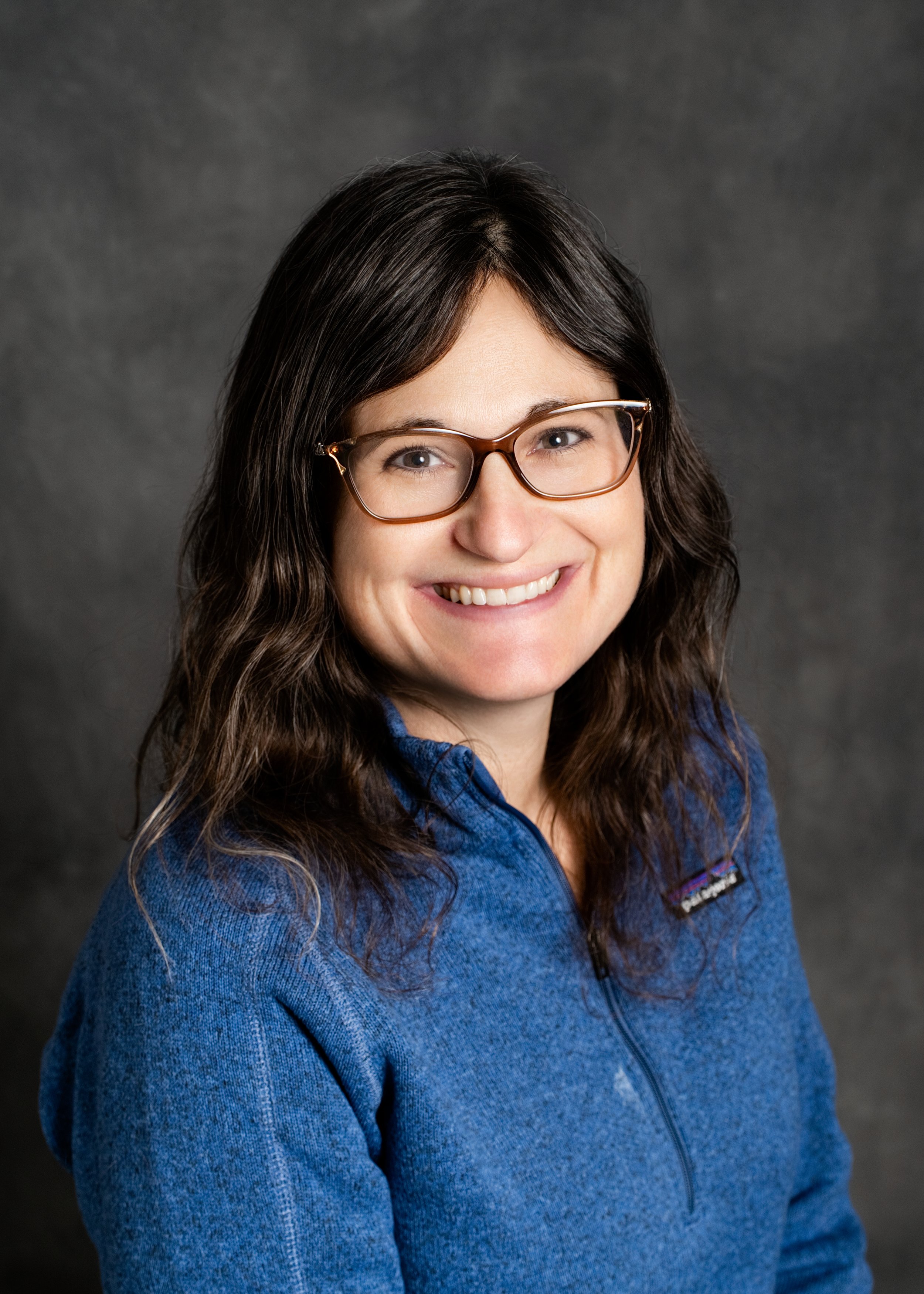 Portrait of Director Ms.Lisa smiling. Woman with long, blonde hair, wearing a pink textured top, against a dark gray background.