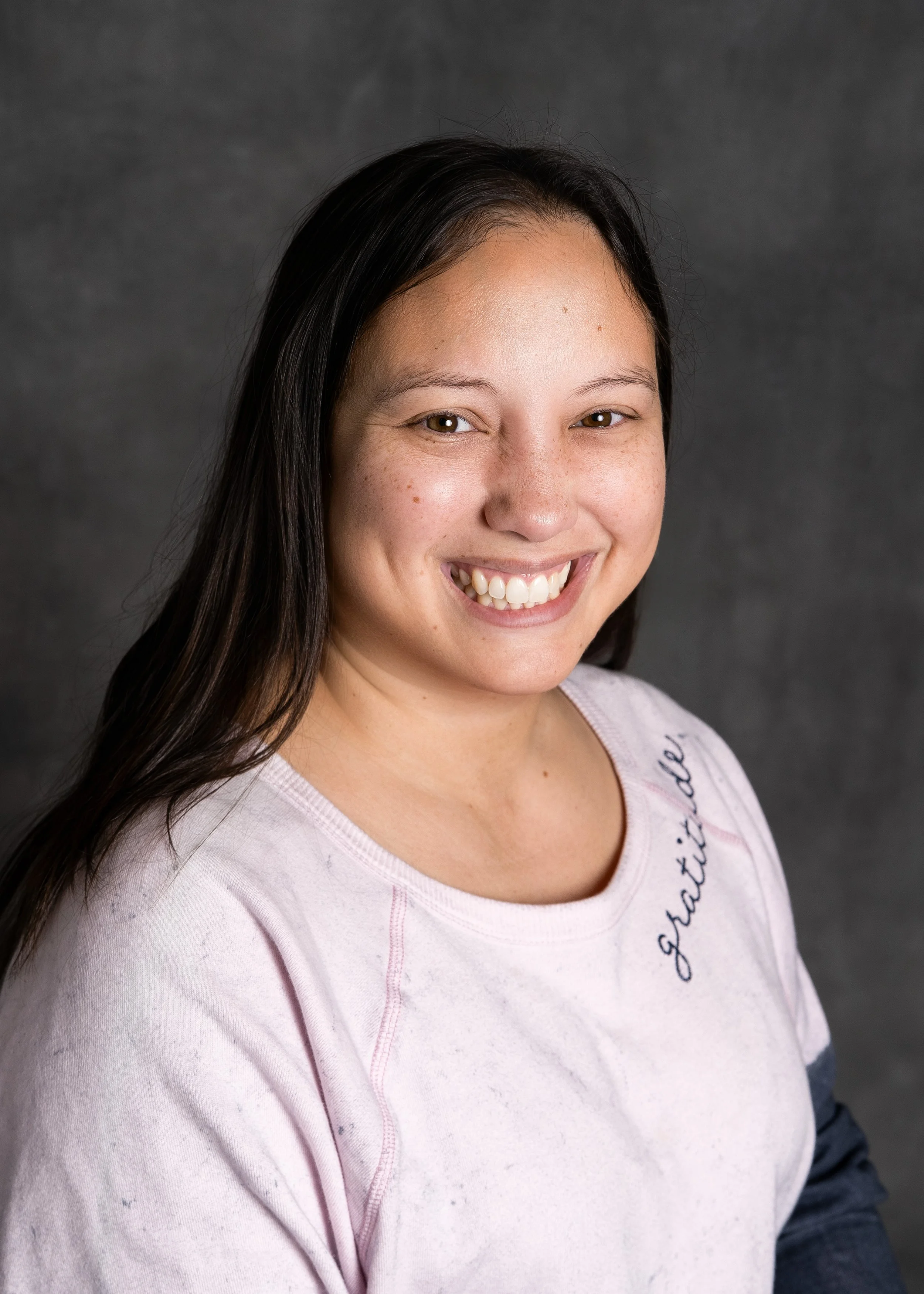 Portrait of teacher Ms.Lauren smiling. Woman with long, blonde hair, wearing a pink textured top, against a dark gray background.