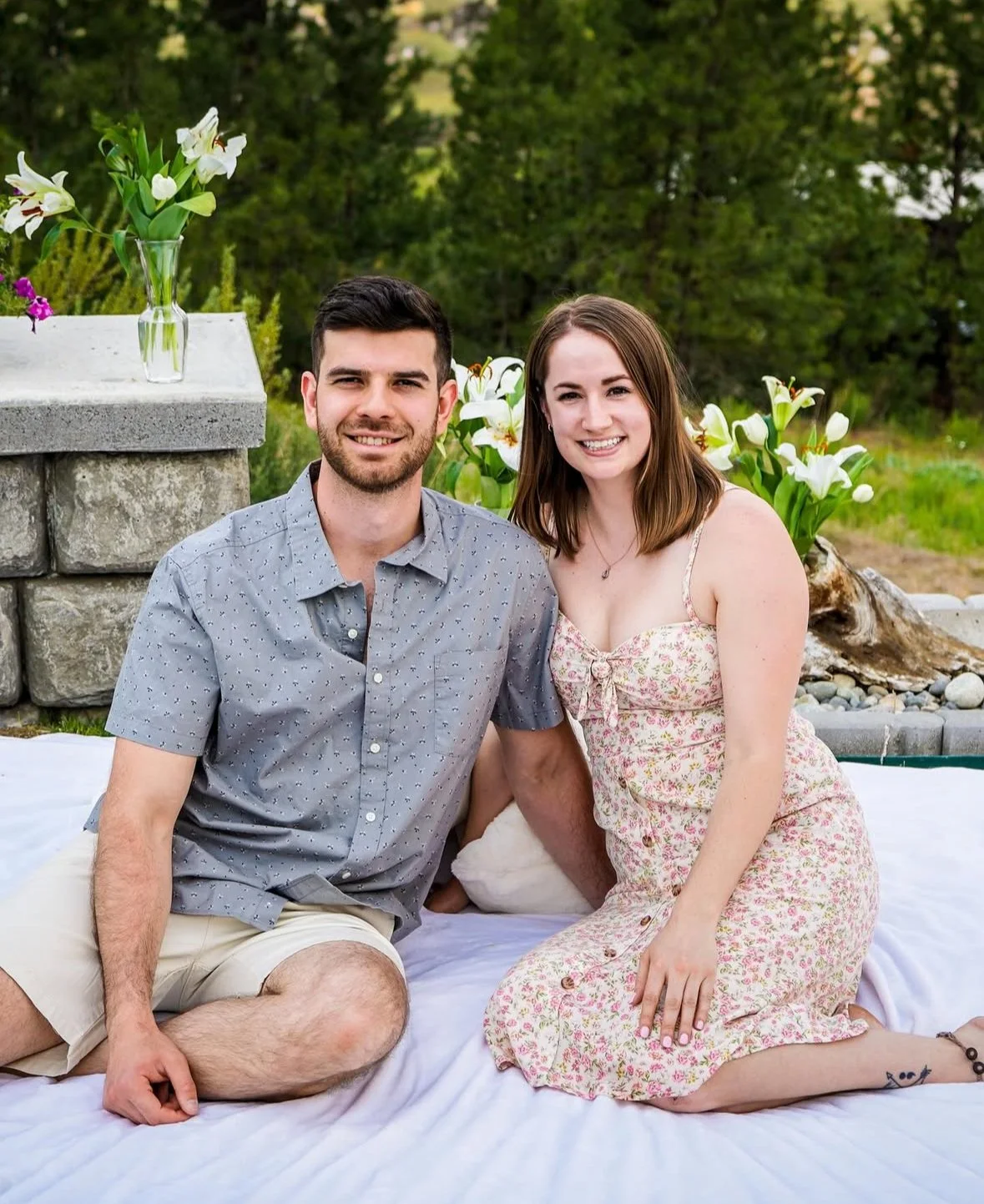A young couple sitting on a white blanket outdoors during daytime, with a stone structure and potted white lilies in the background, smiling at the camera.