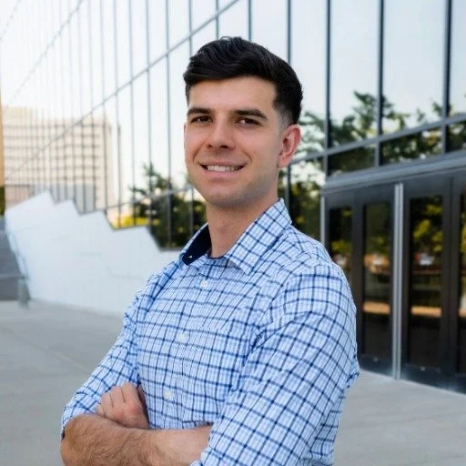 A young man in a blue plaid shirt standing outdoors with his arms crossed, smiling in front of a glass building with reflections and trees in the background.