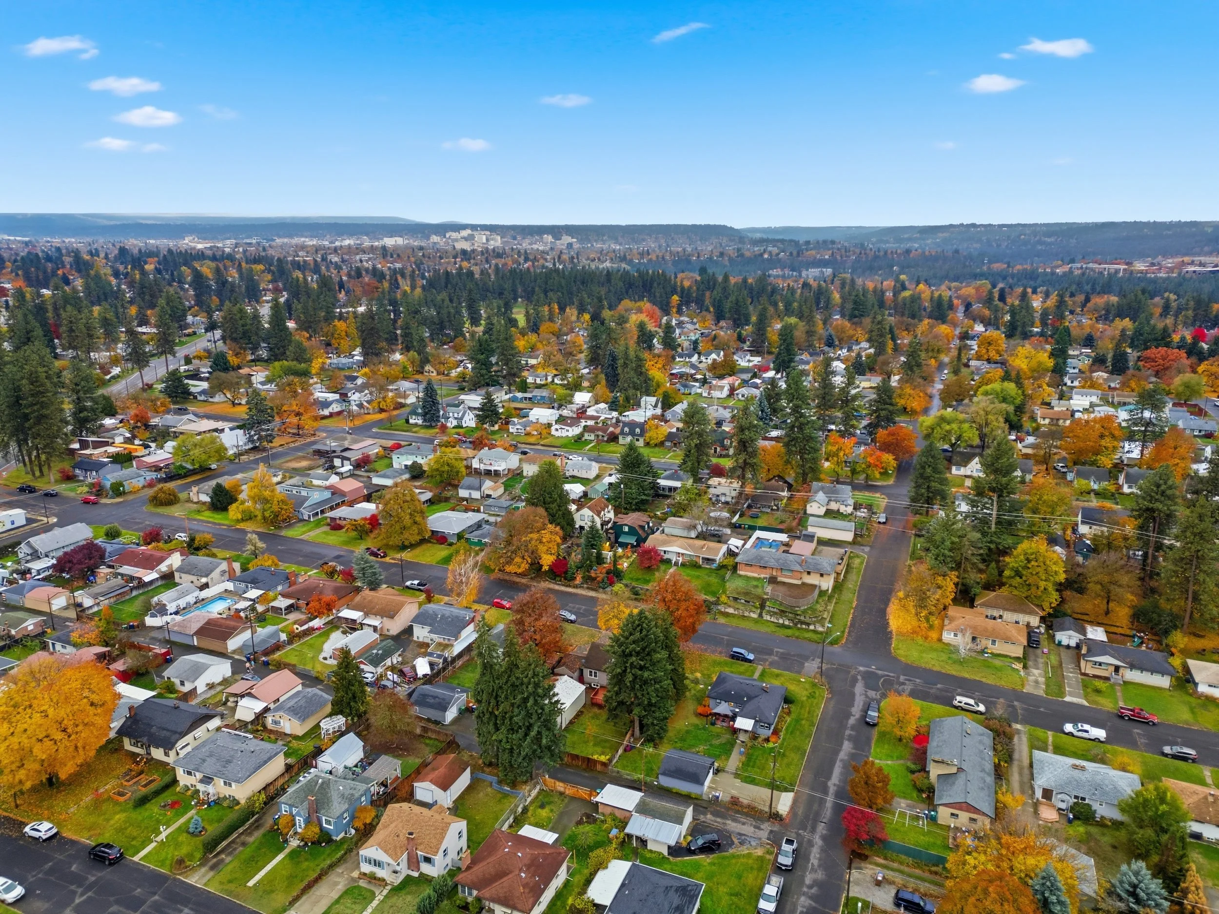 An aerial view of a suburban neighborhood during autumn, with colorful fall foliage on trees, houses, and streets, under a blue sky with scattered clouds.