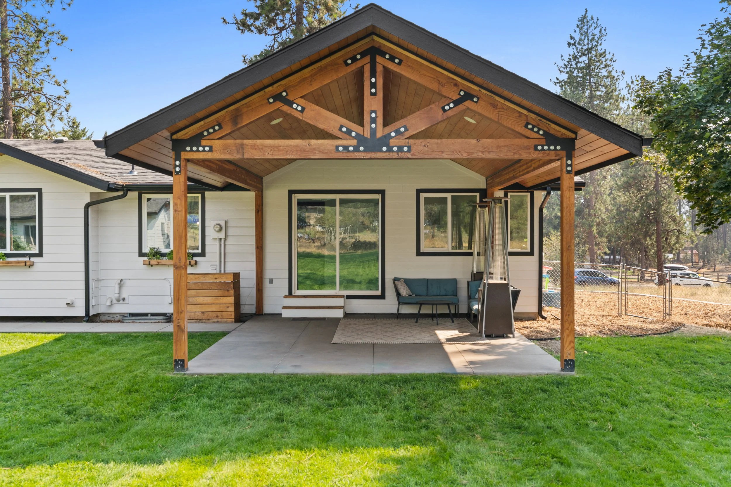 Backyard patio with a white house and a wooden pergola, outdoor seating, and a propane heater, surrounded by green grass and trees in the background.