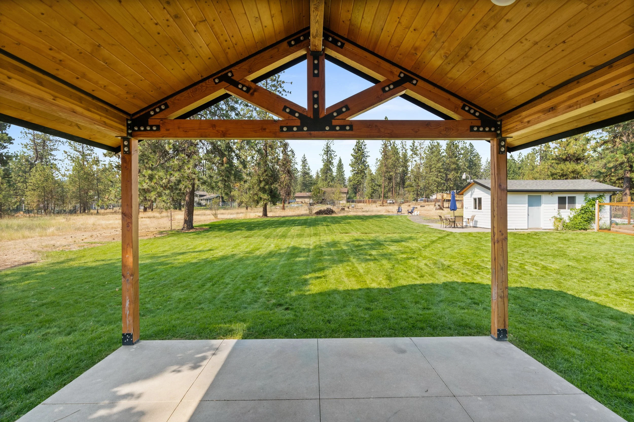View from a covered patio with a wooden ceiling and support beams, looking out onto a green lawn with trees, a small white building, and outdoor furniture in the background.