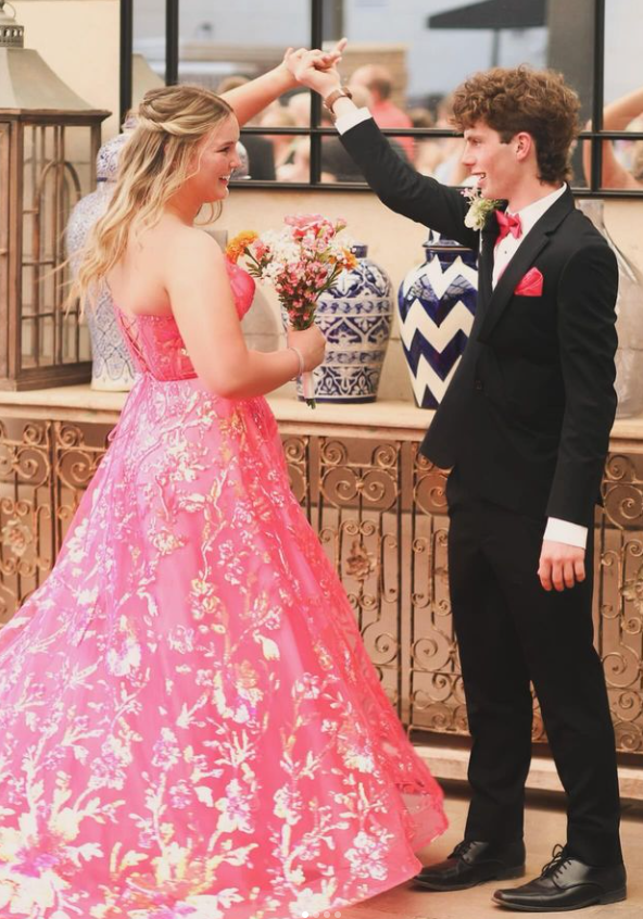 A young couple dancing at a formal event. The girl is wearing a pink floral dress, holding a bouquet, and smiling. The boy is dressed in a black suit with a red pocket square and bow tie. They are twirling in front of a decorative wall with vases and