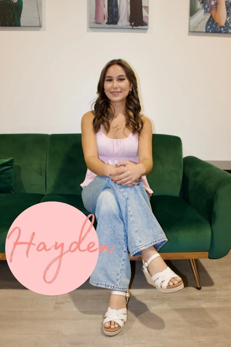 A woman with curly hair and a houndstooth jacket sitting on a dark green sofa, smiling at the camera. The background has a white wall with two photographs and decorative elements including a pink cloud, a pink signature reading 'Avani,' and a pink leaf.