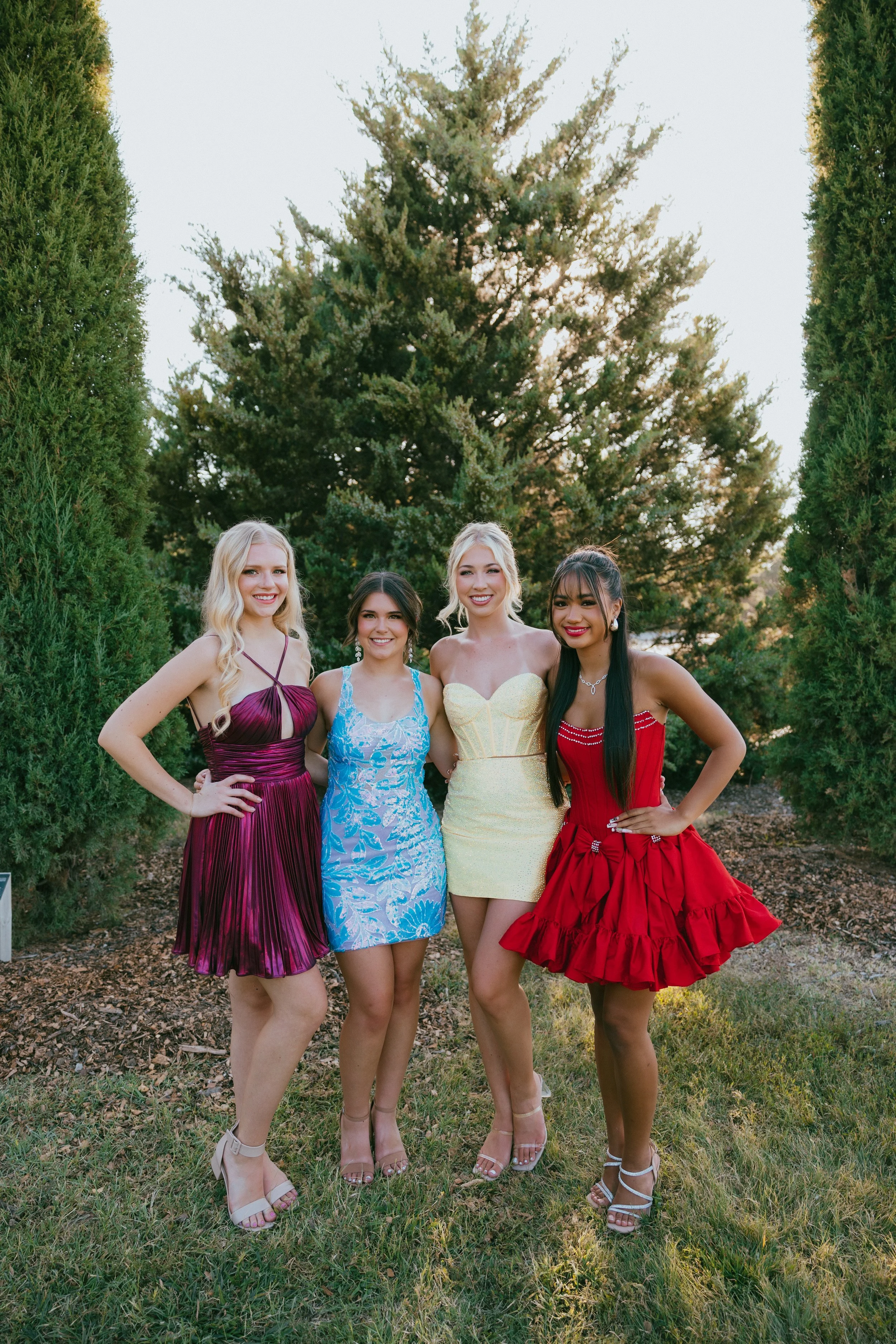 Four young women dressed in colorful dresses standing outdoors on grass, smiling at the camera, with trees in the background.