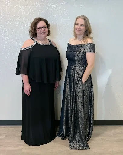 Two women standing side by side in evening dresses, smiling in front of a light-colored decorative wall.