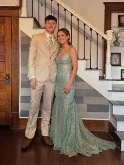 A young man in a beige suit and a young woman in a sparkly green dress standing together indoors in front of a staircase.
