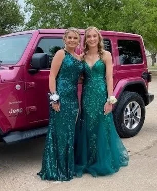 Two young women in emerald prom gowns standing next to a maroon Jeep, smiling at the camera with trees in the background.