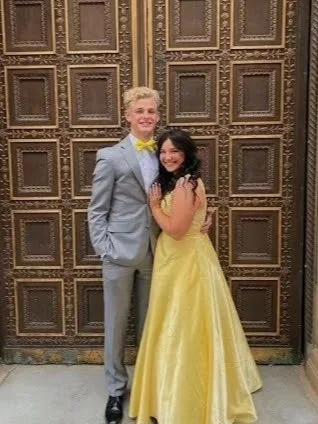 Two people, a young man in a gray suit and a young woman in a yellow ball gown, standing close together and smiling in front of ornate wooden doors.