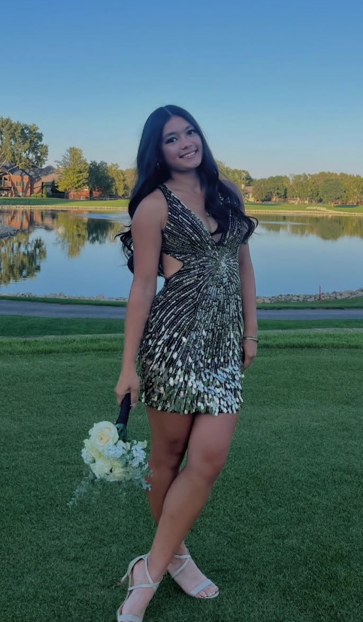 A young woman in a sparkly black and gold Sherri Hill dress holding a white flower bouquet, standing on green grass near a lake with trees and houses in the background during clear weather.