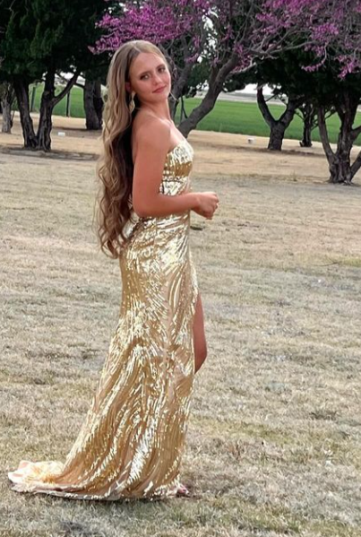 A woman in a gold, strapless, sequined gown standing on a grassy field with trees and pink blossoms in the background.