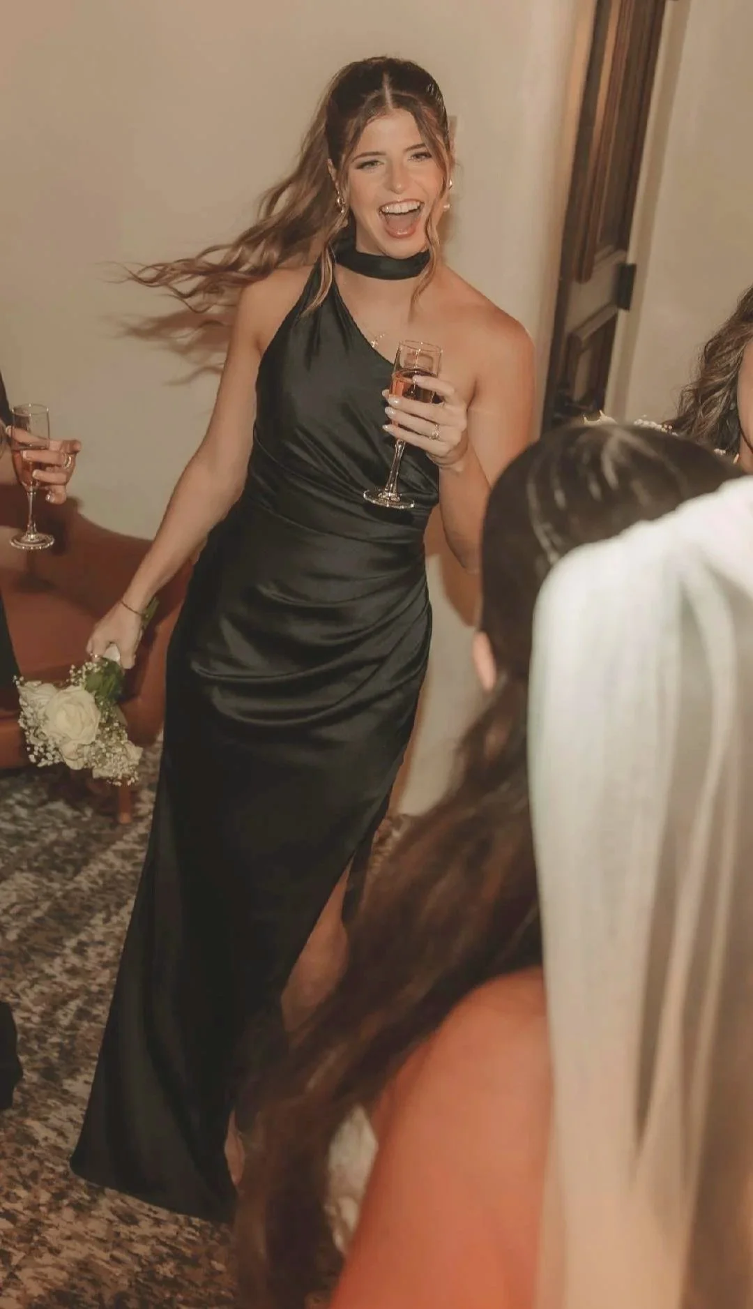 Woman in a black satin gown holding a glass of Rosé wine, smiling and laughing at a celebration or wedding.