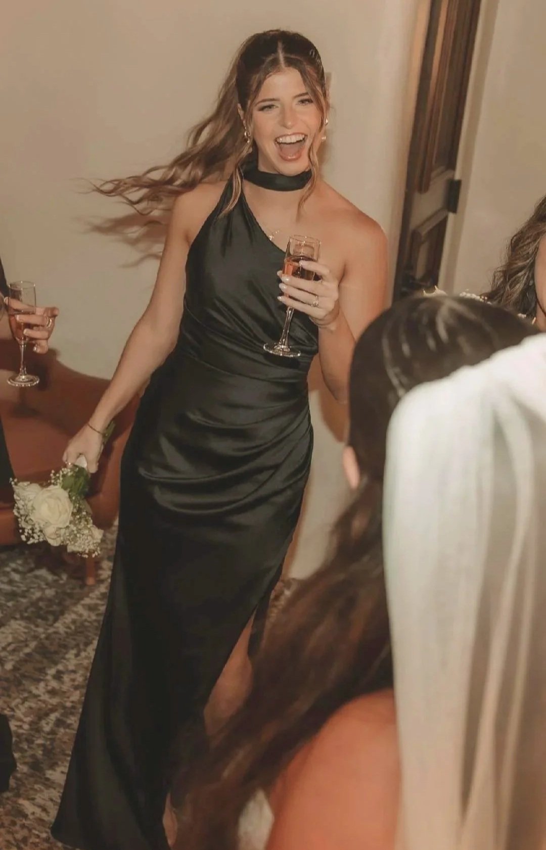 Woman in a black satin gown holding a glass of Rosé wine, smiling and laughing at a celebration or wedding.