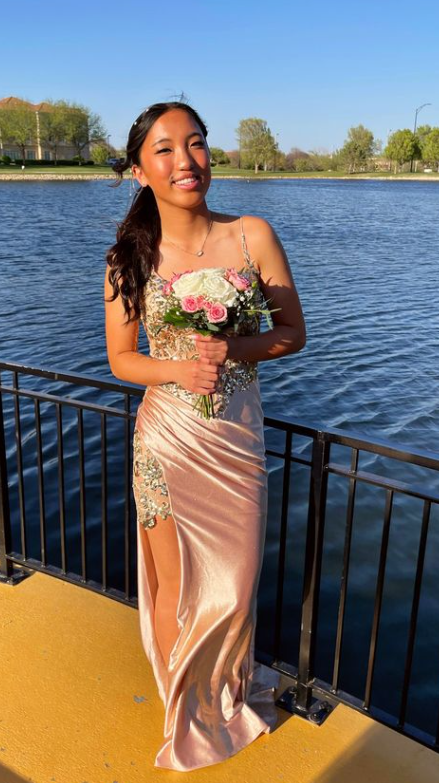 A young woman in a peach satin gown with floral embroidery, holding a bouquet of pink and white flowers, standing by a water body with a railing and trees in the background.