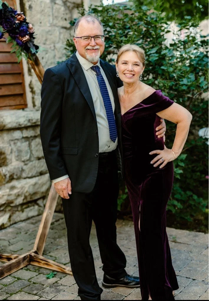 A man in a black suit, white shirt, and blue tie standing next to a woman in a dark purple dress with off-the-shoulder design, both smiling outdoors with greenery and a stone wall in the background.