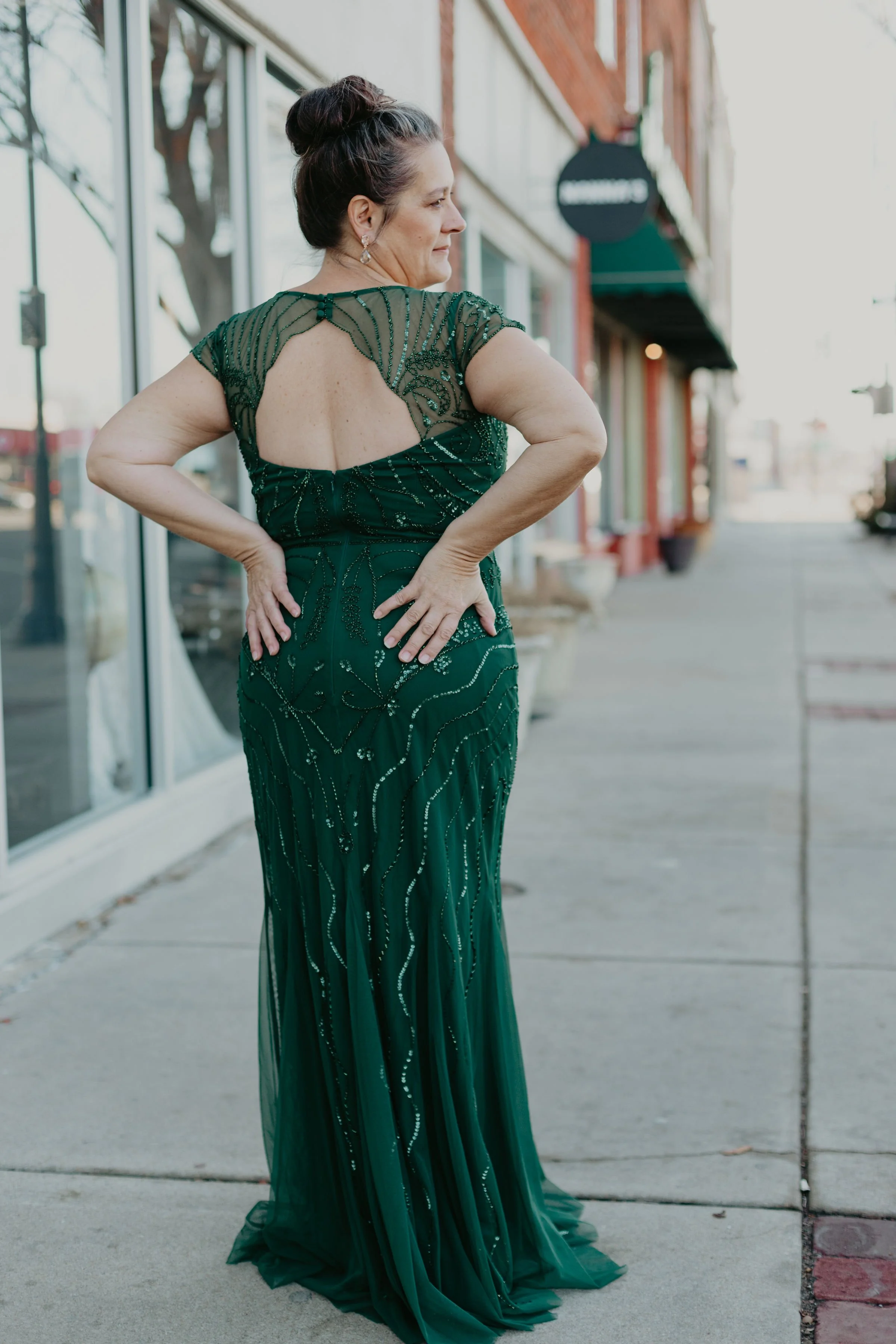 A woman wearing a dark green, embellished evening gown with a keyhole open back, standing on a sidewalk outside a storefront.