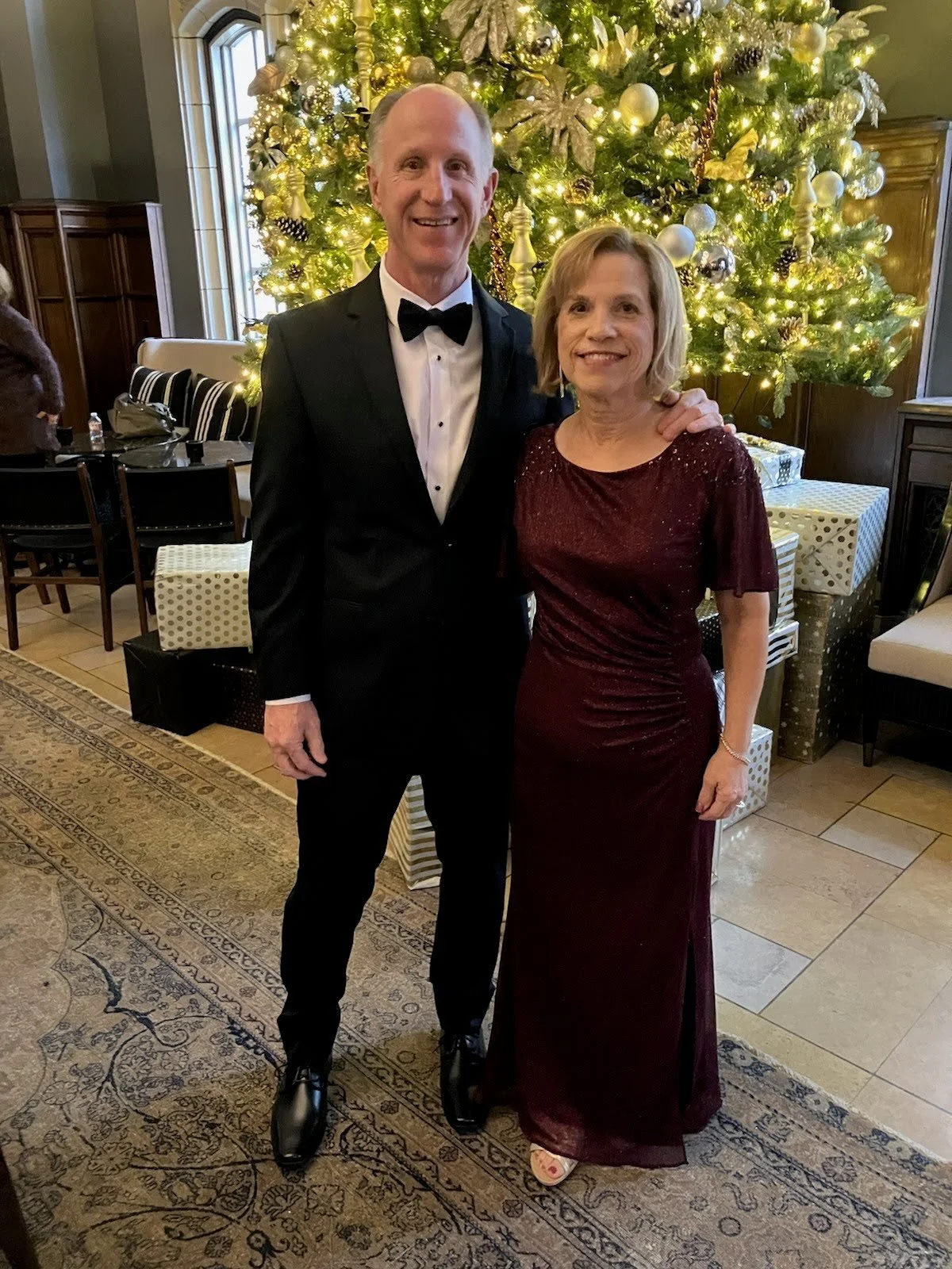 A man in a black tuxedo with a bow tie standing next to a woman in a long burgundy dress, both smiling, in front of a decorated Christmas tree with lights and ornaments, in a warmly lit room.
