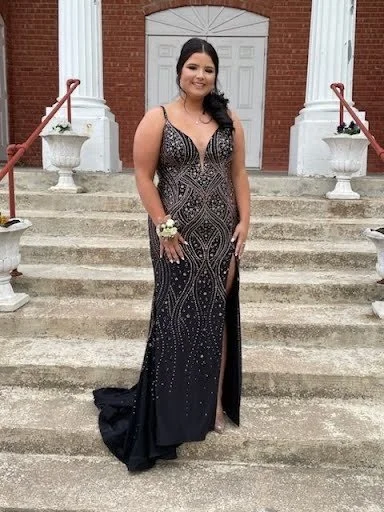 A woman in a long, black, silver-embellished evening gown with a thigh-high slit, standing on the steps in front of a brick and white building, holding a corsage on her wrist, and smiling.