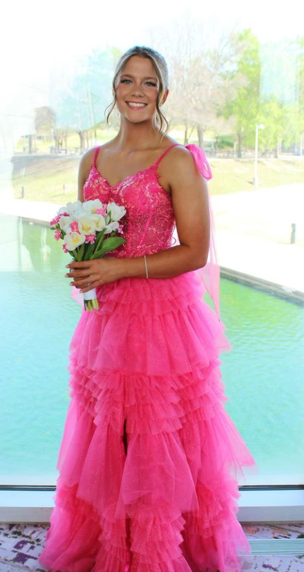 Young woman in a bright pink, ruffled dress holding a bouquet of white and pink flowers, standing indoors near a large window with a view of a pond and trees outside.