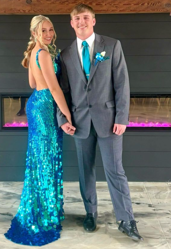 A young man and woman dressed in formal attire, standing indoors against a dark wall with a modern fireplace, holding hands and smiling at the camera.