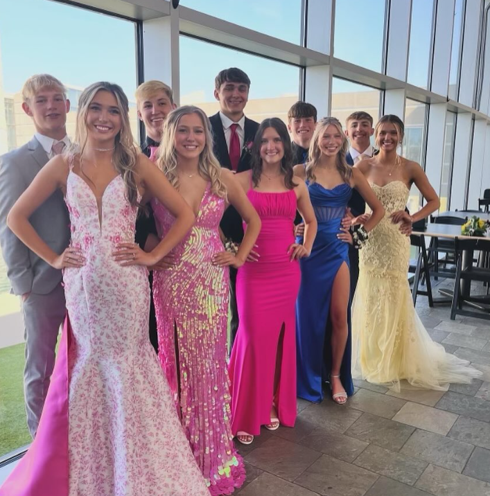 Group of nine young adults in formal attire standing inside a modern building with large windows, smiling for a photo.