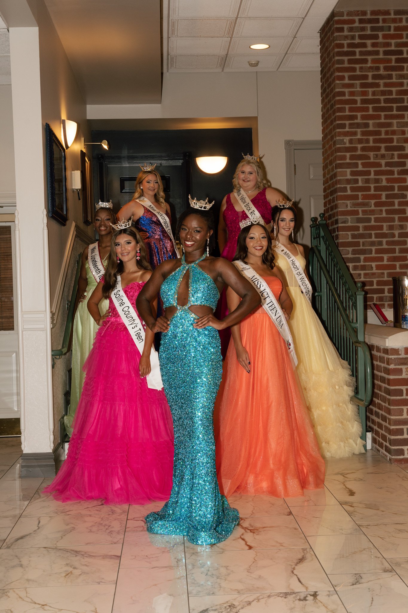 Seven women wearing colorful evening gowns and sashes with crowns, posing together at a formal event indoors.