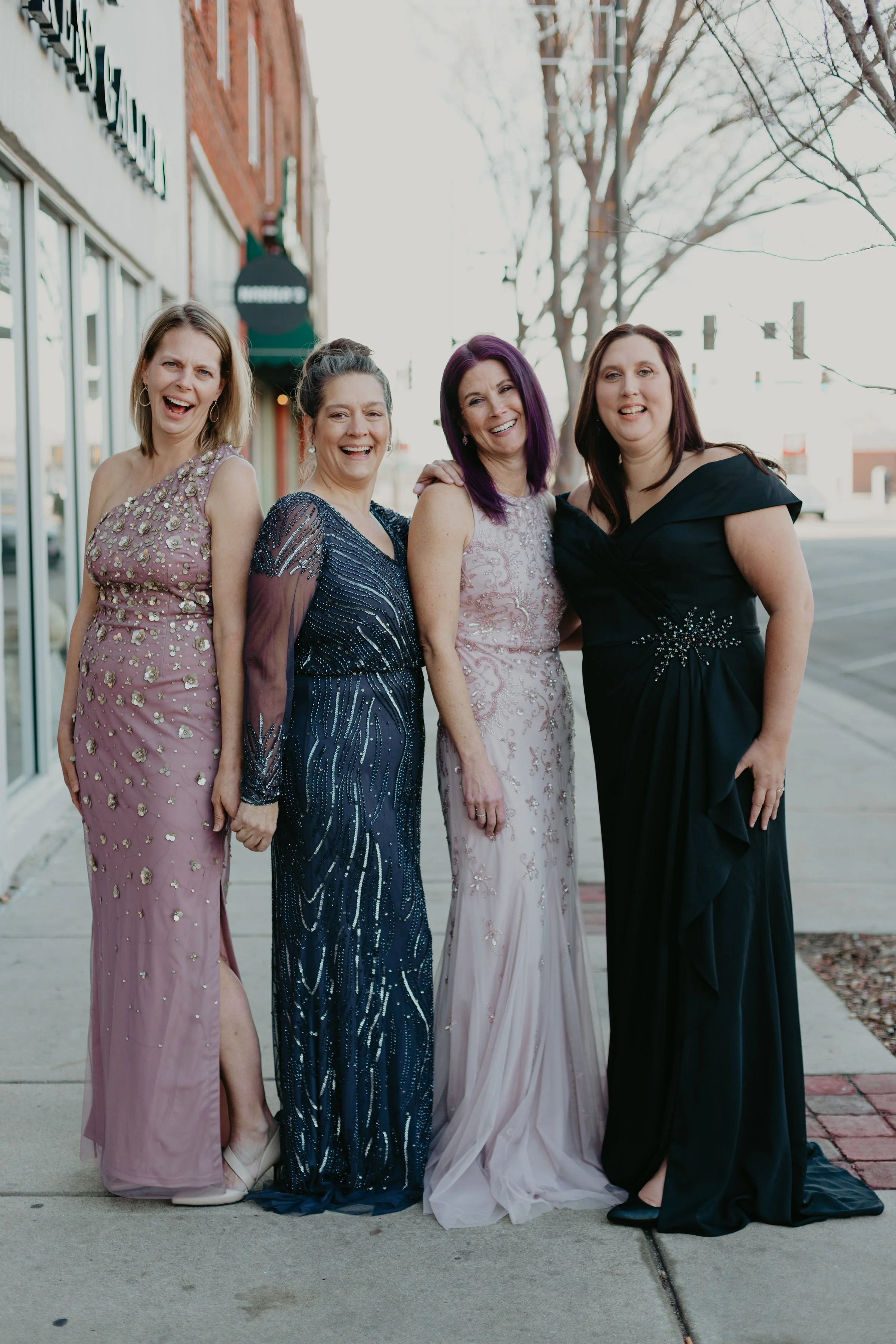 Four women in elegant mother of the bride dresses standing together on a sidewalk, smiling and posing for the photo.