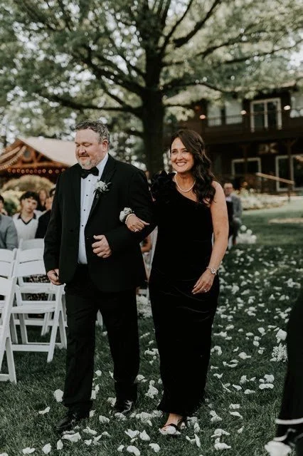 A mother of the bride walking down the aisle with her husband at an outdoor wedding ceremony.