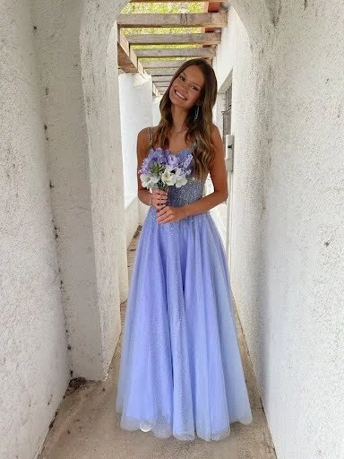 A young woman in a purple and lavender dress holding a bouquet of purple and white flowers, standing in a narrow white-walled corridor.