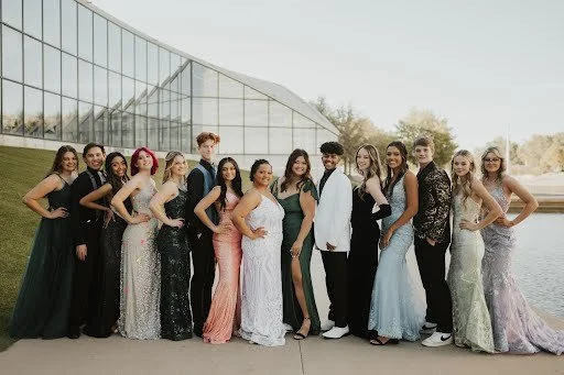Group of young adults dressed in formal attire posing outdoors near a modern glass building and a body of water.