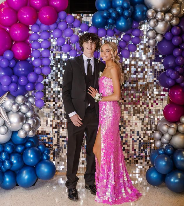A young man in a black suit and a young woman in a pink sequin dress with a thigh slit, posing together at a prom decorated with purple, blue, and silver balloons and a shiny mosaic backdrop.