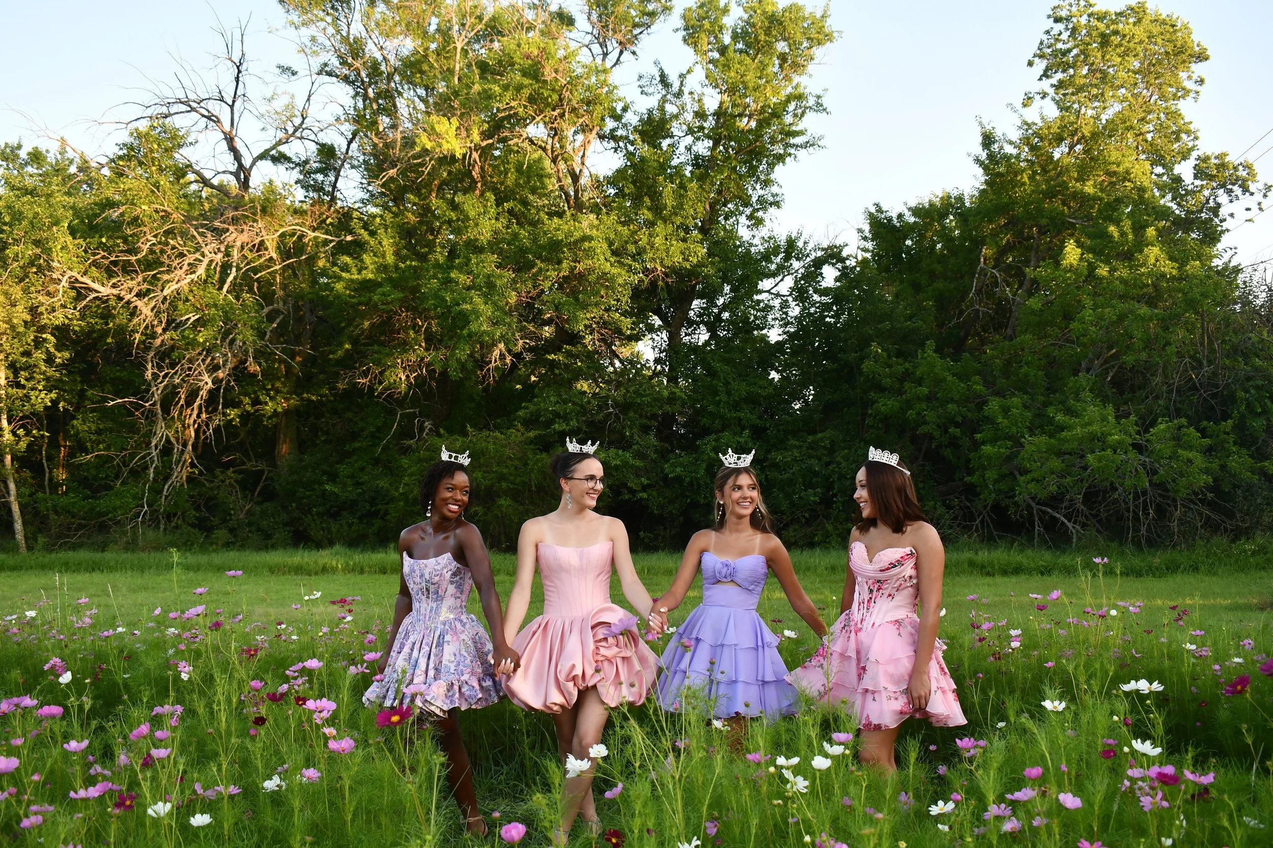 Four young women dressed in colorful dresses standing outdoors on grass, smiling at the camera, with trees in the background.